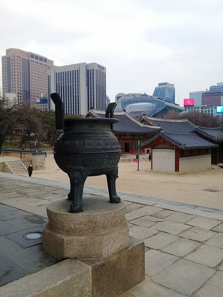 The view from DEOKSUGUNG PALACE, Seoul. In the background there are two 30 stored skyscrapers and another 21st century looking building. In the middle four traditional little palace buildings with black tiles on the roofs. In the foreground an ancient ritual metal black round pot standing on sand-colored stoned ground