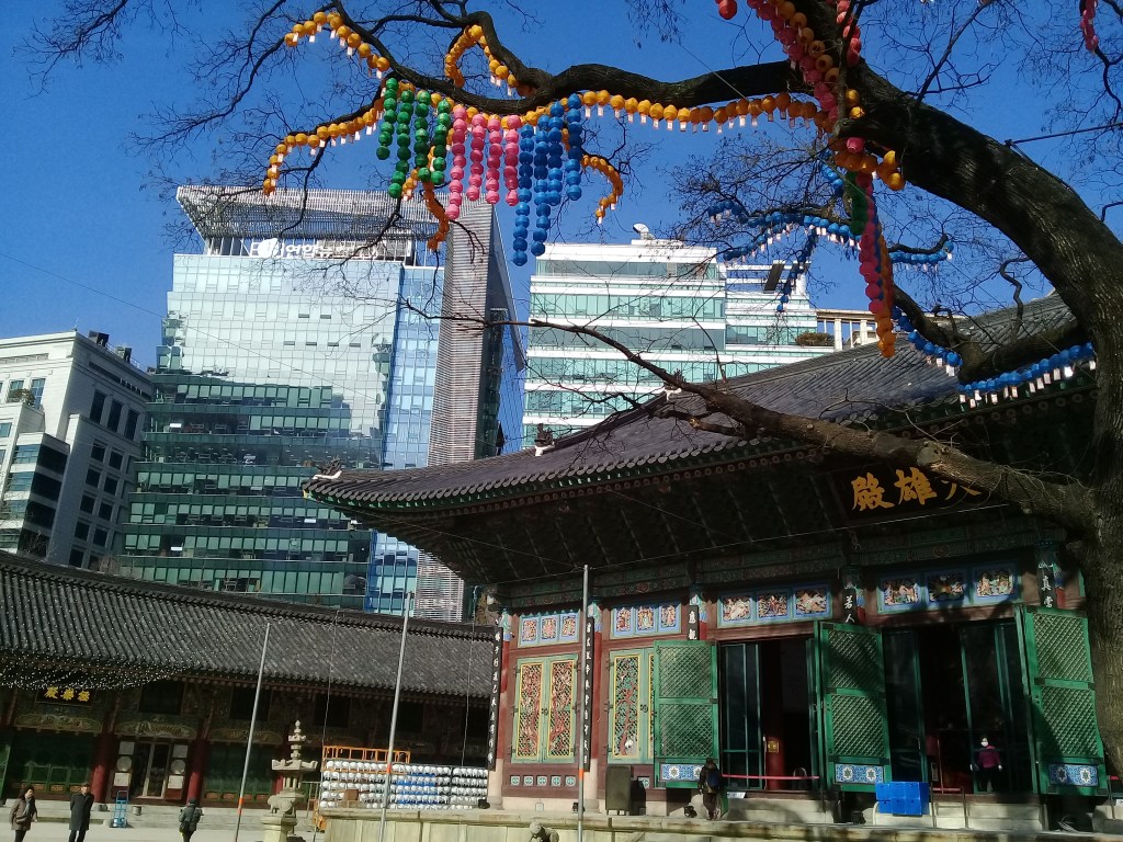 A korean Buddhist temple with a colorfully decorated tree above and two glass tall buildings in the background. 